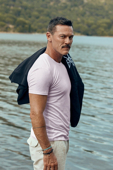Man standing by a lake wearing a pink shirt and white pants with a black jacket over his shoulder.