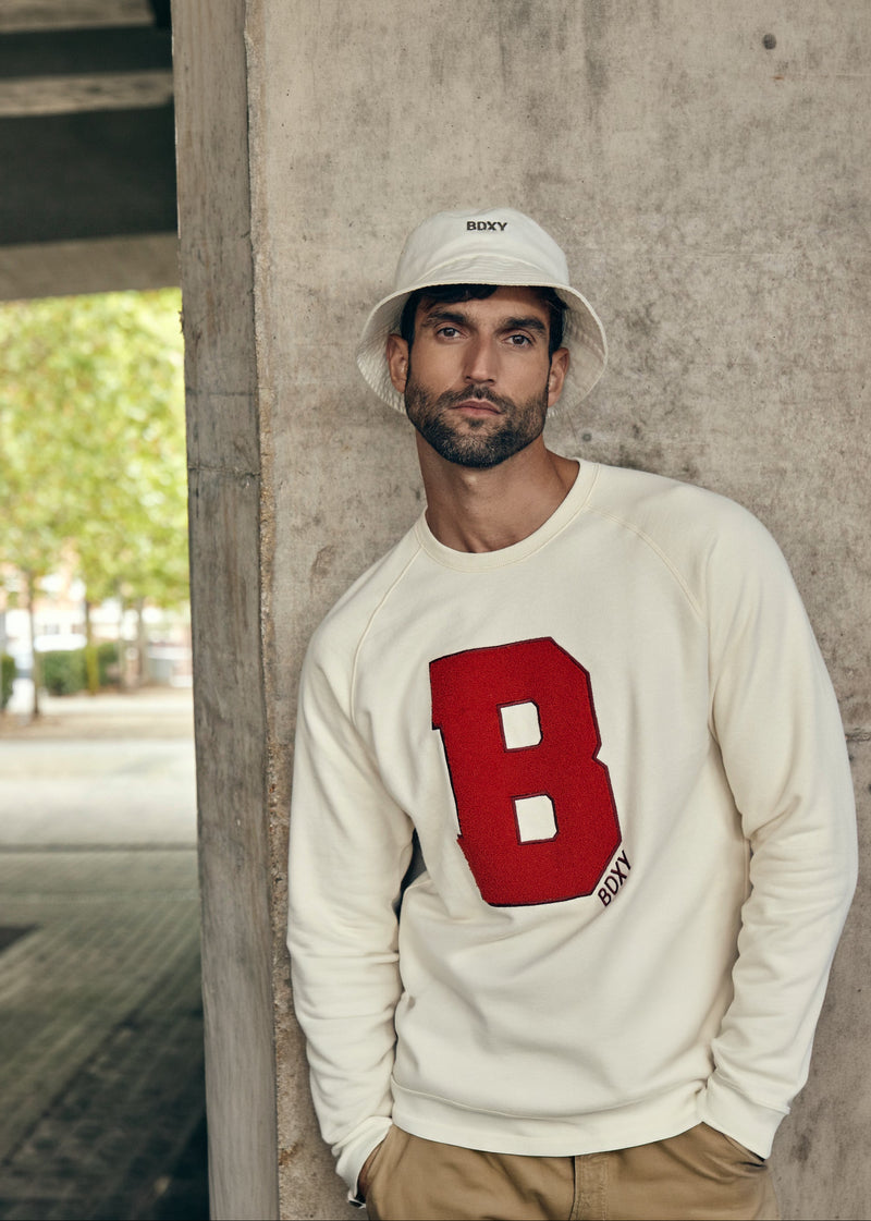 A man wearing a cream-colored crewneck sweatshirt with a large red block letter 'B' on the front, khaki pants, and a white bucket hat with 'SENKY' printed on it, standing against a concrete pillar.