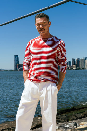 A man wearing a red and white striped long-sleeve shirt and white trousers stands outdoors by the water with a city skyline in the background.