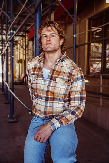 A man wearing a brown, beige, and white plaid flannel shirt over a white undershirt and blue jeans, posing on a city sidewalk under scaffolding.