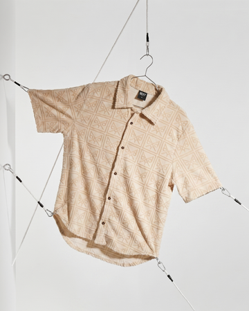 A short-sleeve button-up shirt with a beige and white geometric diamond pattern, hanging from a black hanger suspended by cables inside a clear display case.