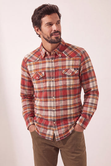 A man wearing a red, orange, and beige plaid western-style button-down shirt with brown pants, standing against a plain light background.